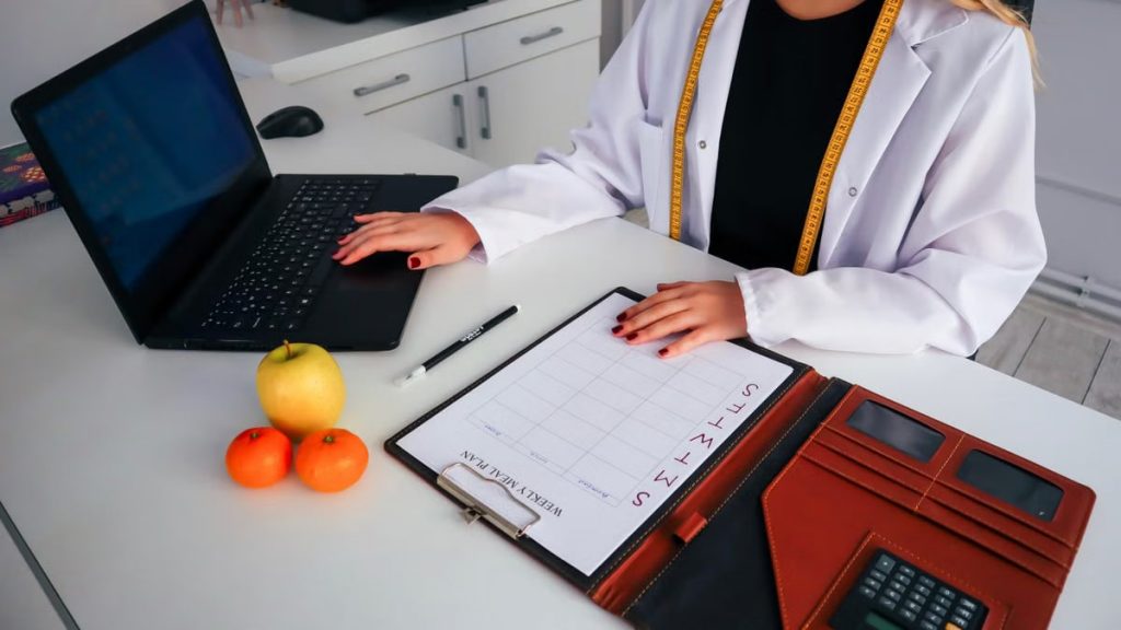 Dietitian working on meal plan with laptop, fruits, and calendar for health consultation.